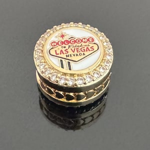 May include: A gold-toned Las Vegas souvenir charm. The charm features the words "Welcome to Fabulous Las Vegas Nevada" in red and white, surrounded by a ring of clear crystals. The base has a heart-shaped design.