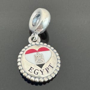 May include: A silver charm with a round pendant featuring the Egyptian flag colors of red, white, and black in a heart shape. The word "EGYPT" is inscribed in silver around the edge of the pendant. The charm has a loop for attaching to a bracelet or necklace.