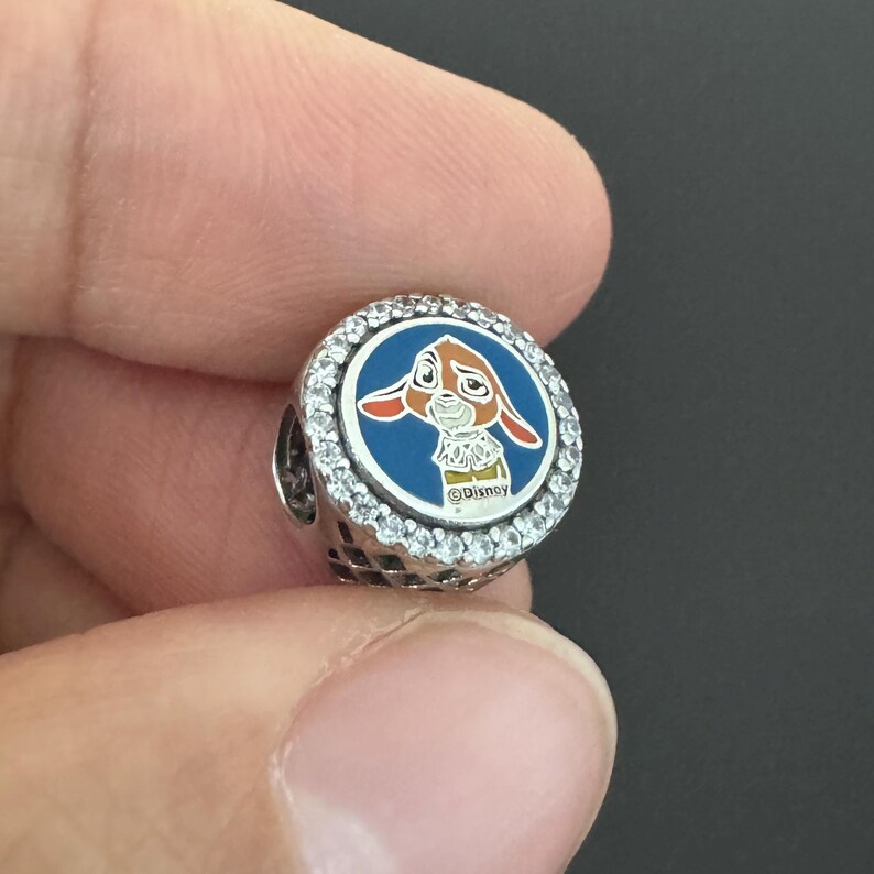 Puede incluir: Un charm de plata con un dise&ntilde;o de conejo de dibujos animados. El conejo es marr&oacute;n y blanco, sobre un fondo azul, y rodeado de cristales transparentes. El charm tiene el logotipo de Disney.