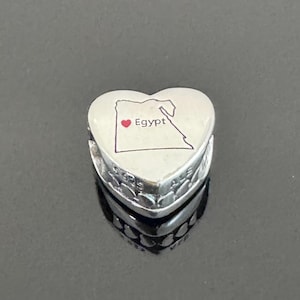 May include: A heart-shaped charm with a map of Egypt and the word "Egypt" in a white background. The charm is silver with a decorative edge. The map is outlined in black with a red heart.
