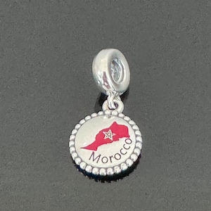 May include: Silver charm with a red and white enamel outline of Morocco with a star and the word "Morocco" written below.