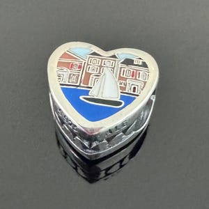 May include: Heart-shaped silver charm with a nautical scene. The charm features a sailboat on blue water, with buildings in the background. The charm is detailed with enamel and has the text "925 ALE" on the side.