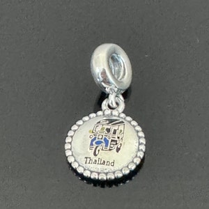 May include: Silver charm with a blue and yellow tuk-tuk on a white background. The charm has a beaded border and says "Thailand" at the bottom.