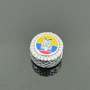 May include: A silver-coloured, round charm with a detailed design. The charm features the Ecuadorian flag in the centre, surrounded by a ring of clear crystals. The charm has a textured, patterned base.