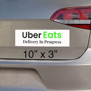 Peut inclure: Arrière d'une voiture argentée avec un autocollant Uber Eats. L'autocollant blanc porte l'inscription verte et noire "Uber Eats Delivery In Progress". En dessous, le texte indique "10" x 3" (25,4 cm x 7,6 cm).