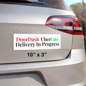 Może przedstawiać: Biały prostokątny magnes z napisem "DoorDash UberEats Delivery In Progress" w kolorze czerwonym, zielonym i czarnym. Magnes ma wymiary 25,4 cm na 7,6 cm i jest przymocowany do tyłu srebrnego samochodu.
