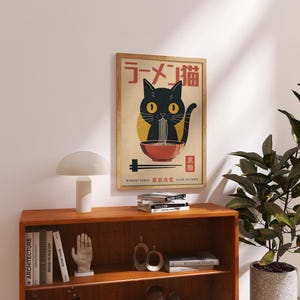 May include: A framed poster featuring a black cat eating ramen noodles. The poster has Japanese text at the top and bottom, with a black cat illustration in the center. The poster is displayed above a wooden shelf with books and a lamp.