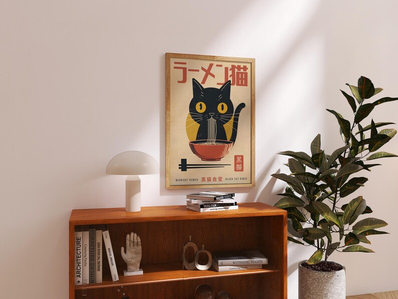 May include: A framed poster featuring a black cat eating ramen noodles. The poster has Japanese text at the top and bottom, with a black cat illustration in the center. The poster is displayed above a wooden shelf with books and a lamp.