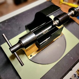 May include: A black metal tool with a T-shaped handle, mounted on a green surface with a grey circular disc. The tool has a gold-coloured strip and a black cylindrical component.