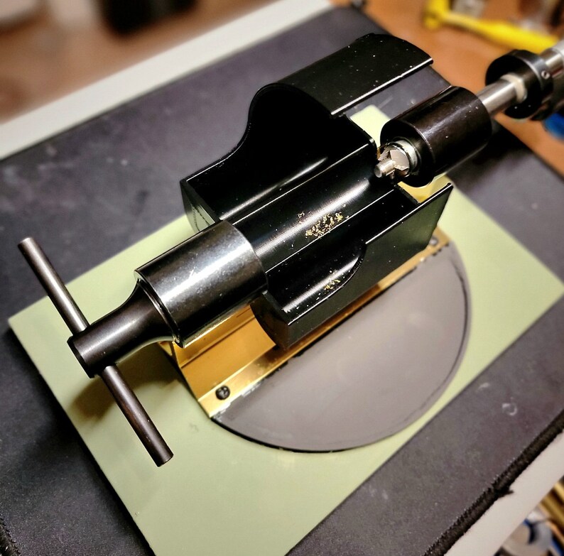 May include: A black metal tool with a T-shaped handle, mounted on a green surface with a grey circular disc. The tool has a gold-coloured strip and a black cylindrical component.