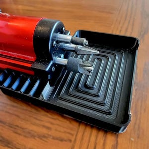 Puede incluir: Una herramienta rotativa roja y negra con varias brocas y una bandeja negra. La herramienta tiene un cuerpo rojo y una cabeza negra con múltiples accesorios. La bandeja negra tiene un diseño acanalado.