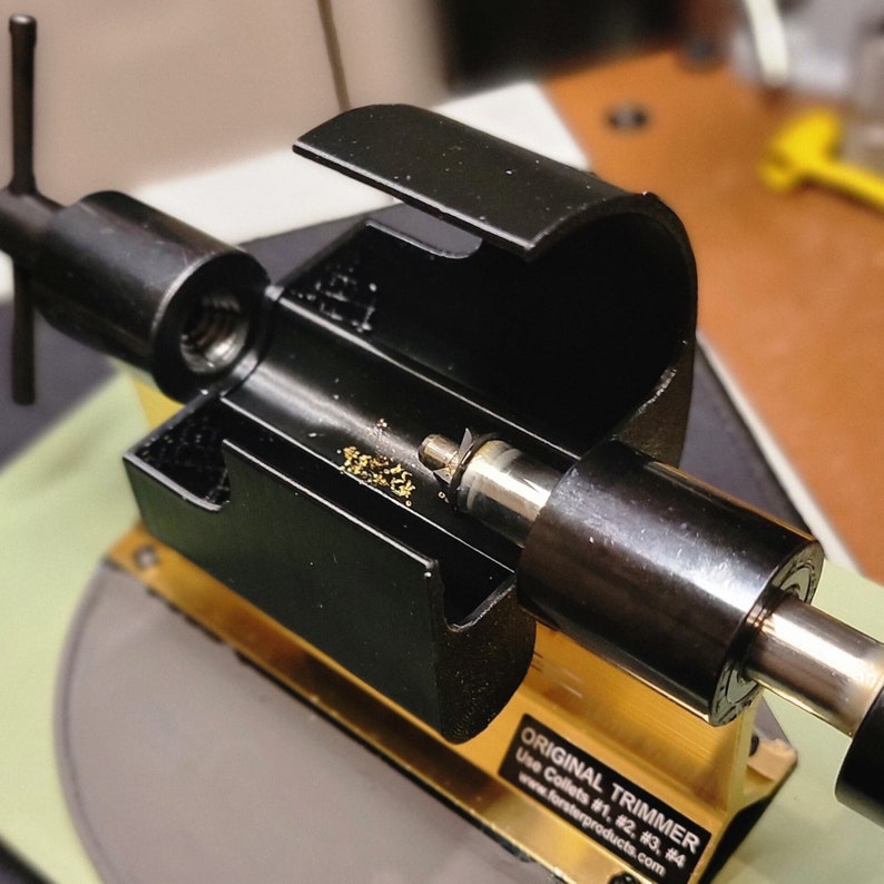 May include: A black metal tool with a gold base. The tool has a handle with a T-shaped grip and a long, cylindrical barrel. The barrel has a small, round opening at one end and a larger, round opening at the other end. The tool is labelled "ORIGINAL TRIMMER" and "Use Collets #1, #2, #3, #4" with the website address "www.forsterproducts.com".