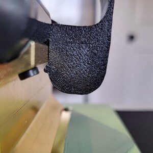 May include: Close-up of a black plastic 3D printed part with a textured surface. The part is attached to a gold metal object.