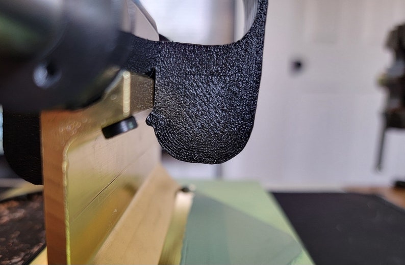 May include: Close-up of a black plastic 3D printed part with a textured surface. The part is attached to a gold metal object.