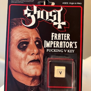May include: A novelty collectible item featuring a white computer keyboard key with the letter V, encased in black plastic packaging. The card displays a portrait of a person with dark eye makeup and the text Ghost, Frater Imperator's Fucking V Key.
