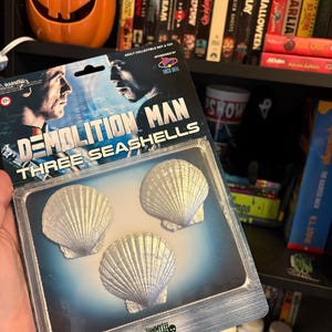 Puede incluir: Un set de colección Demolition Man Three Seashells en su empaque blíster original. La tarjeta presenta el título de la película, la marca Taco Bell y tres réplicas de conchas marinas texturizadas de color plateado en una burbuja de plástico.