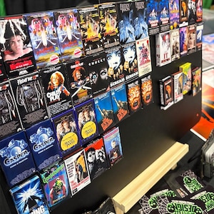 May include: A collection of miniature VHS tapes featuring classic horror movies like "Friday the 13th," "Child's Play," and "Scream." The tapes are displayed on a black surface, showcasing the colorful movie covers. The collection includes titles like "Casper," "Goosebumps," and "Halloween."