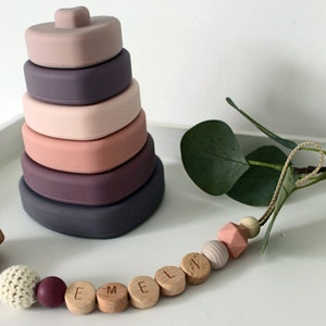 May include: A pink and purple silicone stacking toy with a heart-shaped top. A wooden teething toy with a clip and beads that spell "EMMA" is also pictured.
