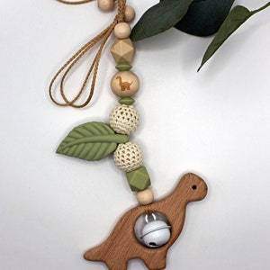 May include: A wooden dinosaur teether with a bell inside, attached to a string of wooden and crocheted beads, green silicone leaves, and a small wooden dinosaur charm.