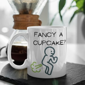 May include: White ceramic coffee mug with a black outline of a person with a green fart cloud. The mug has the text "FANCY A CUPCAKE?" printed on it.