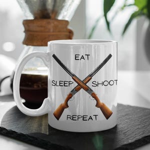 May include: White ceramic coffee mug with a black and brown graphic of two crossed shotguns. The text "Eat Sleep Shoot Repeat" is printed on the mug.