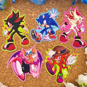 May include: Five colorful stickers featuring characters from the Sonic the Hedgehog franchise. The stickers are on a brown corkboard background. The characters are Sonic, Shadow, Shadow the Hedgehog, Rouge the Bat, and Knuckles.
