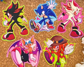 Könnte beinhalten: Fünf bunte Aufkleber mit Charakteren aus dem Sonic the Hedgehog-Franchise. Die Aufkleber befinden sich auf einem braunen Korkuntergrund. Die Charaktere sind Sonic, Shadow, Shadow the Hedgehog, Rouge the Bat und Knuckles.