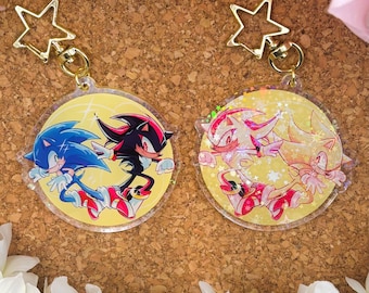 Könnte beinhalten: Zwei runde, transparente, holografische Schlüsselanhänger mit Charakteren aus der Sonic the Hedgehog Videospiel-Reihe. Der erste Schlüsselanhänger zeigt Sonic und Shadow, während der zweite Shadow und Silver zeigt. Beide Schlüsselanhänger haben einen goldenen Stern-Anhänger.