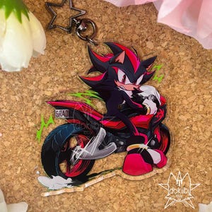 May include: An acrylic keychain featuring a black and red hedgehog character riding a red and black motorcycle. The character has red markings and white gloves. The motorcycle has a license plate that reads SHAD. A black star-shaped metal clasp is attached.
