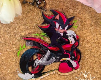 May include: An acrylic keyring featuring a black and red hedgehog character riding a red and black motorcycle. The character has red markings and white gloves. The motorcycle has a number plate that reads SHAD. A black star-shaped metal clasp is attached.