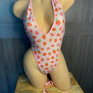 Puede incluir: Un bañador rosa con cuello halter y tanga a juego con estampado de fresas. El bañador tiene un escote bajo y está ribeteado en rosa. El diseño de fresas incluye fresas rojas y naranjas sobre un fondo blanco.