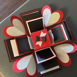May include: A red and black explosion box with silver glitter hearts. The box has multiple layers and is decorated with a red bow and a silver star.