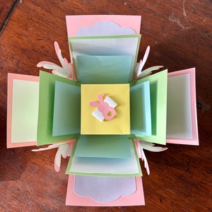 May include: A multi-layered paper craft box in pastel colors. The box features pink, green, blue, and yellow paper elements. A small bunny cutout with a bow is centered on the top layer. The box is open, revealing multiple layers of paper.