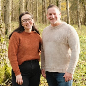 May include: Two people wearing knitted sweaters, standing in a forest. The woman is wearing a rust-colored sweater and the man is wearing a light beige sweater.