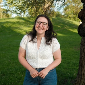 Peut inclure: Une femme aux cheveux foncés et aux lunettes sourit en portant un cardigan en crochet blanc avec un devant boutonné et un jean bleu.