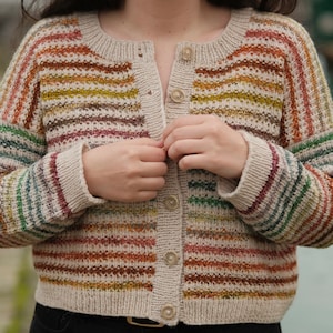 Peut inclure: Un cardigan rayé avec une fermeture boutonnée. Le cardigan est fait d'un fil doux et texturé dans des tons de brun, beige et vert.
