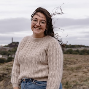 Peut inclure: Une femme aux longs cheveux bruns portant des lunettes sourit à la caméra. Elle porte un pull en tricot beige clair et un jean bleu. L'arrière-plan est une vue floue d'une ville.