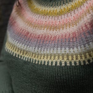 May include: A close-up of a green sweater with a high, ribbed collar. The yoke features horizontal stripes in shades of yellow, pink, and gray. The sweater is made of a knitted fabric.