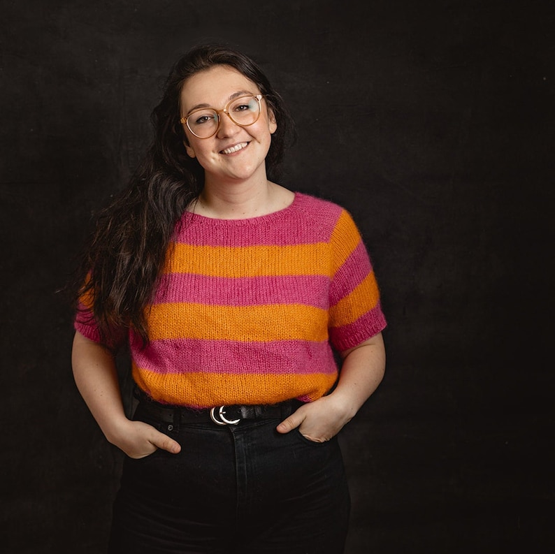May include: A woman wearing a pink and orange striped sweater with a black belt and black jeans. She is smiling and looking at the camera.