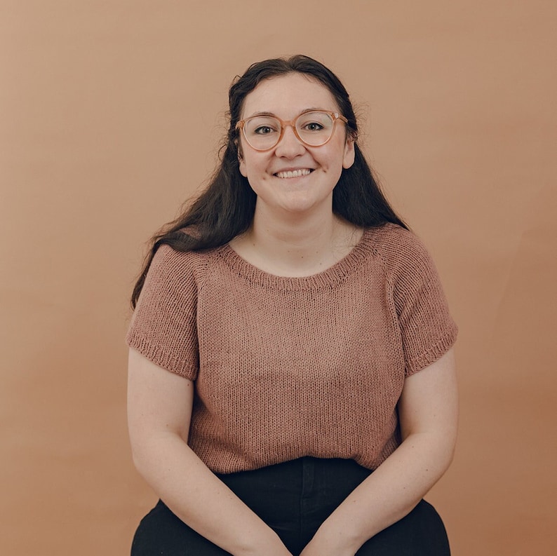 May include: A woman with long dark hair is wearing a mauve knitted sweater with short sleeves and black pants. She is smiling and wearing glasses.