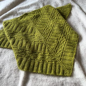 May include: A green knitted shawl with a geometric pattern. The shawl is folded and laid on a white fuzzy surface.