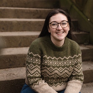 Op de afbeelding: Een vrouw met een bril glimlacht terwijl ze op een stenen trap zit. Ze draagt een groene en beige trui met een patroon.