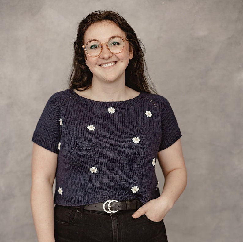 May include: A woman wearing a navy blue sweater with white embroidered flowers. She is wearing a black belt and black jeans. She is smiling and looking at the camera.