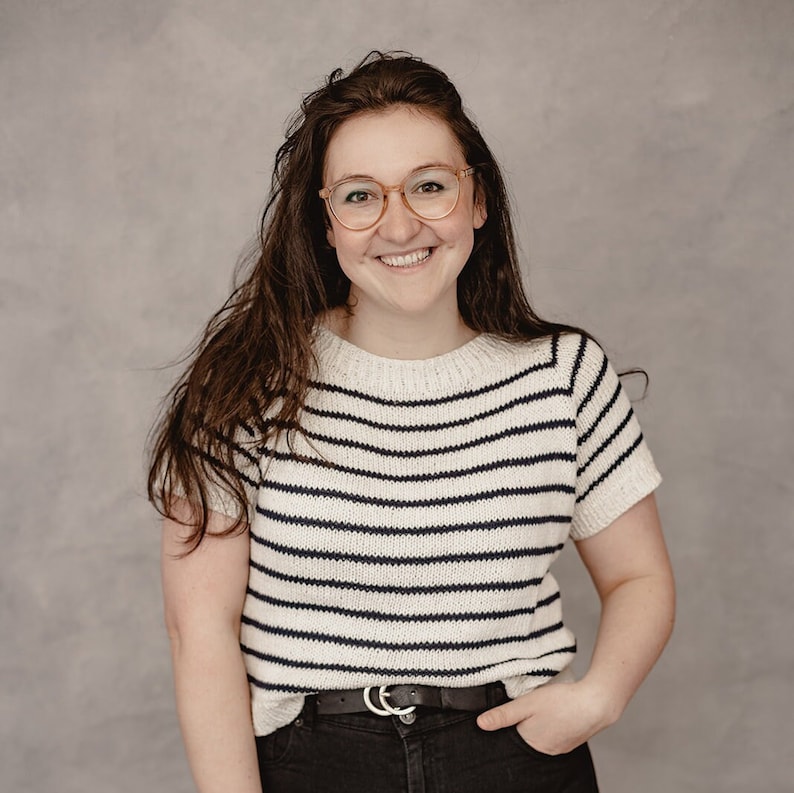 May include: A woman with long brown hair is wearing a white and navy striped shirt with a black belt. She is smiling and looking at the camera.