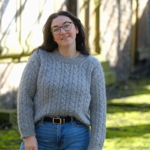 Peut inclure: Une femme portant un pull gris en tricot torsadé avec une ceinture noire et un jean bleu. Elle porte des lunettes et sourit.