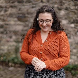 Peut inclure: Une femme portant un cardigan en crochet orange vif avec un devant boutonné. Elle sourit et regarde vers le bas. Le cardigan a un motif en dentelle et est porté sur une robe à pois noirs et blancs.