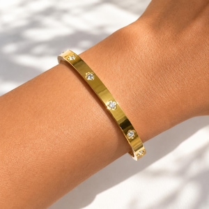 Puede incluir: Un brazalete rígido de tono dorado pulido que presenta una serie de pequeñas piedras transparentes de corte redondo, espaciadas uniformemente a lo largo de la banda metálica. El brazalete tiene una superficie lisa y reflectante.