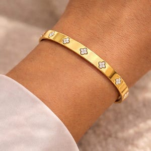 May include: A gold-colored bangle bracelet with a series of four-petal flower designs, each set with small, sparkling gemstones. The bracelet is worn on a wrist, and the background is a soft, neutral color.
