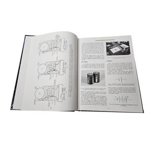 May include: An open book with black covers, displaying detailed diagrams and text. The pages feature illustrations of circuits, batteries, and electrical components. Two Procell batteries are shown. The book is likely a technical manual or textbook.