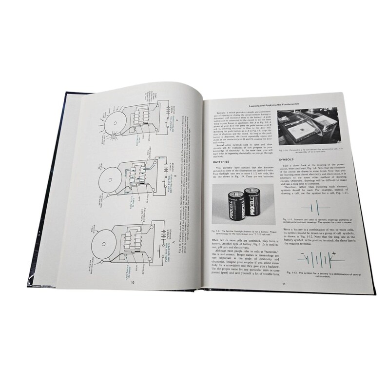 May include: An open book with black covers, displaying detailed diagrams and text. The pages feature illustrations of circuits, batteries, and electrical components. Two Procell batteries are shown. The book is likely a technical manual or textbook.